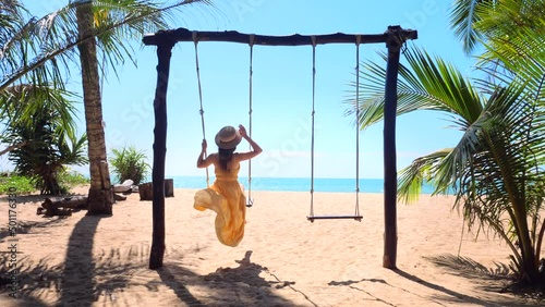 Slow motion of unrecognizable travel woman on wooden swing on tropical sandy beach with blue sea and palm trees. Female traveler on Phuket island, Thailand. Vacation and summer holidays concept. 