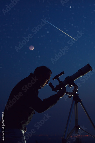 Silhouette of a man, telescope, stars, planets and shooting star under the night sky.