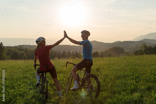 Road cyclists, man and woman, stopping at a grassy hill to celebrate the race achievement with high five gesture, at sunset light.