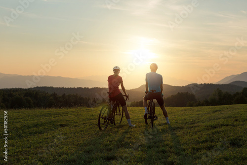 Professional cyclist couple taking a break on a green meadow and watching amazing mountain sunset, aerial shot.