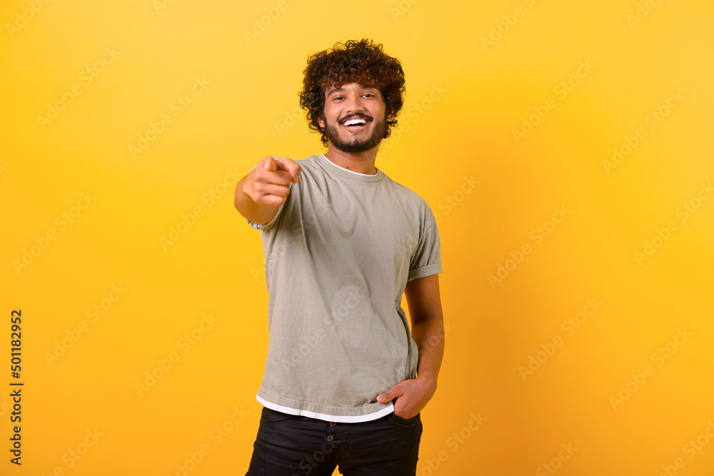 Smiling young indian man with curly hairstyle pointing finger at you standing isolated on yellow background, cheerful teenage guy indicating at the camera Smiling young indian man with curly hairstyle pointing finger at you standing isolated on yellow background, cheerful teenage guy indicating at the camera
