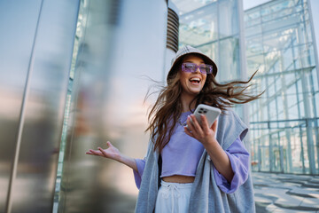 Excited young smiling woman holding and using mobile phone. Cool stylish girl at outside