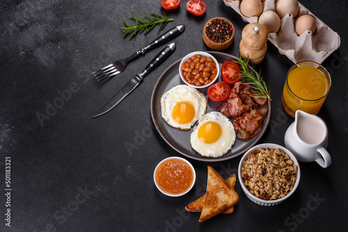 Fototapeta Naklejka Na Ścianę i Meble -  A delicious nutritious healthy breakfast with fried eggs, bacon, beans, a glass of juice, oat cookies, milk and jam