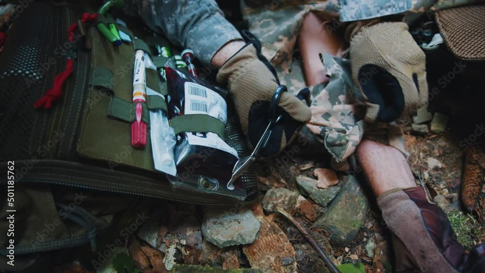 Close up hands military medic cuts the clothes of a wounded soldier with scissors to examine the body. Soldiers lying on ground. Blood around Close up hands military medic cuts the clothes of a wounded soldier with scissors to examine the body. Soldiers lying on ground. Blood around