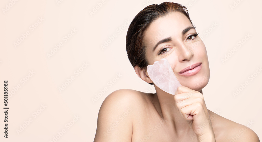 Beauty and skincare concept with a beautiful woman doing skin care routine with gua sha tool. Naked brunette girl using guasha to lymphatic drainage and reduce wrinkles. Beige background banner Beauty and skincare concept with a beautiful woman doing skin care routine with gua sha tool. Naked brunette girl using guasha to lymphatic drainage and reduce wrinkles. Beige background banner