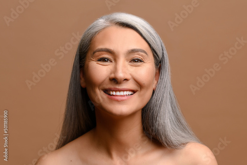 Happy beautiful middle-aged grey haired Asian woman posing in studio on brown background. Cheerful mature korean lady with fresh and hydrated skin looks at the camera and laughing