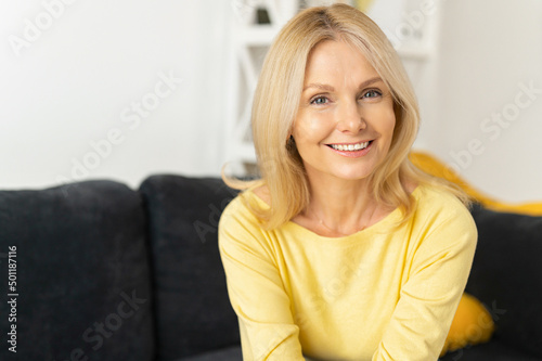 Senior woman in home interior. Charming mature caucasian lady looking at the camera and laughing, resting at the sofa. Serene female spends weekend at home