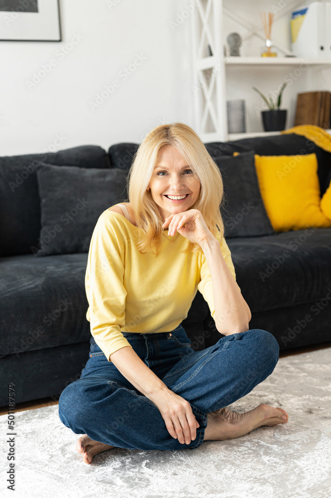 Serene carefree middle-aged woman with long hair sitting near the sofa in modern apartment, senior female resting at home, daydreaming, laughing Serene carefree middle-aged woman with long hair sitting near the sofa in modern apartment, senior female resting at home, daydreaming, laughing
