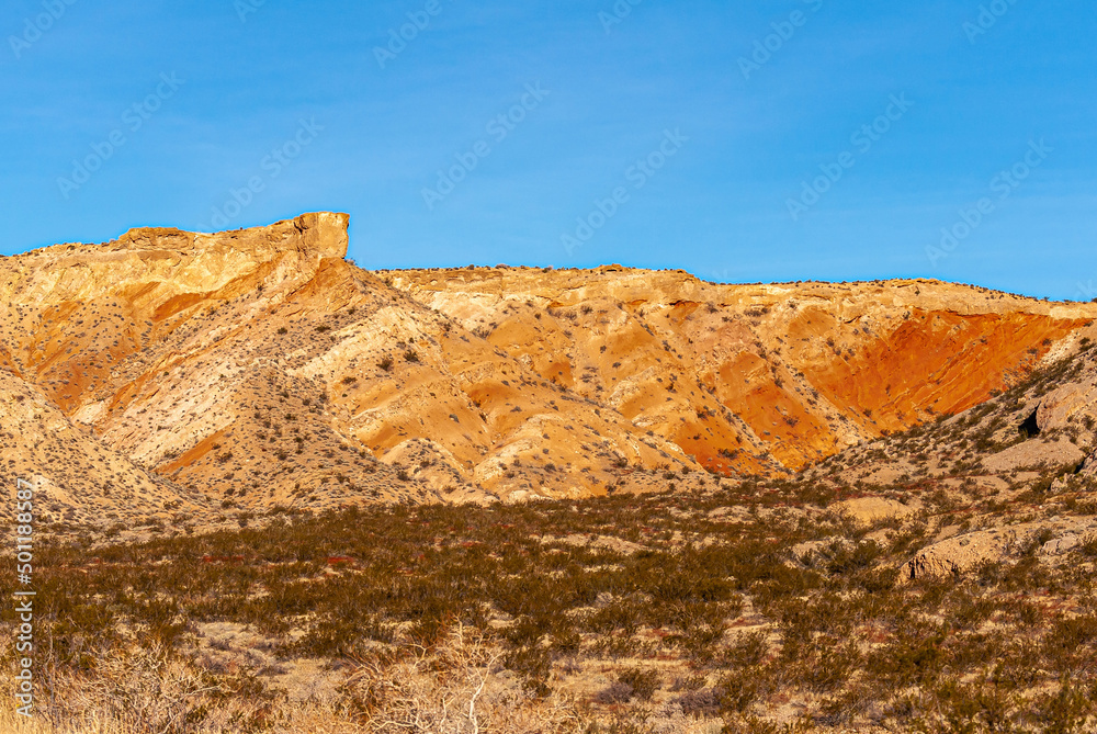 Fototapeta premium Overton, Nevada, USA - February 24, 2010: Valley of Fire. Orange-yellow mountain range forms a blet between blue sky and dry desert floor with brown shrubs.
