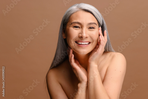 Enchanting topless middle aged Asian woman looks at the camera and laughing happily, isolated on brown background, charming korean lady with grey hair holding face gently. Skin care and self love