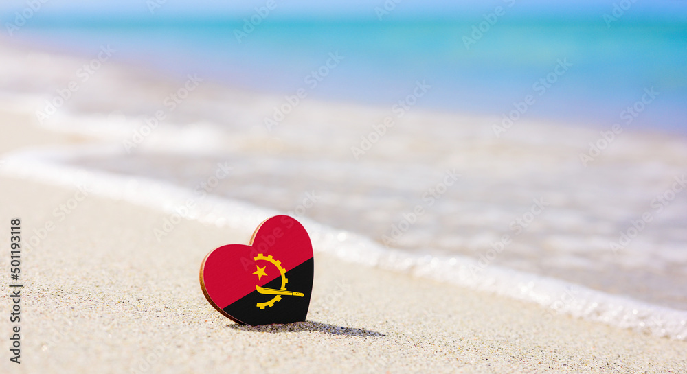 Obraz premium Flag of Angola in the shape of a heart on a sandy beach. The concept of the best vacation in Angola resorts