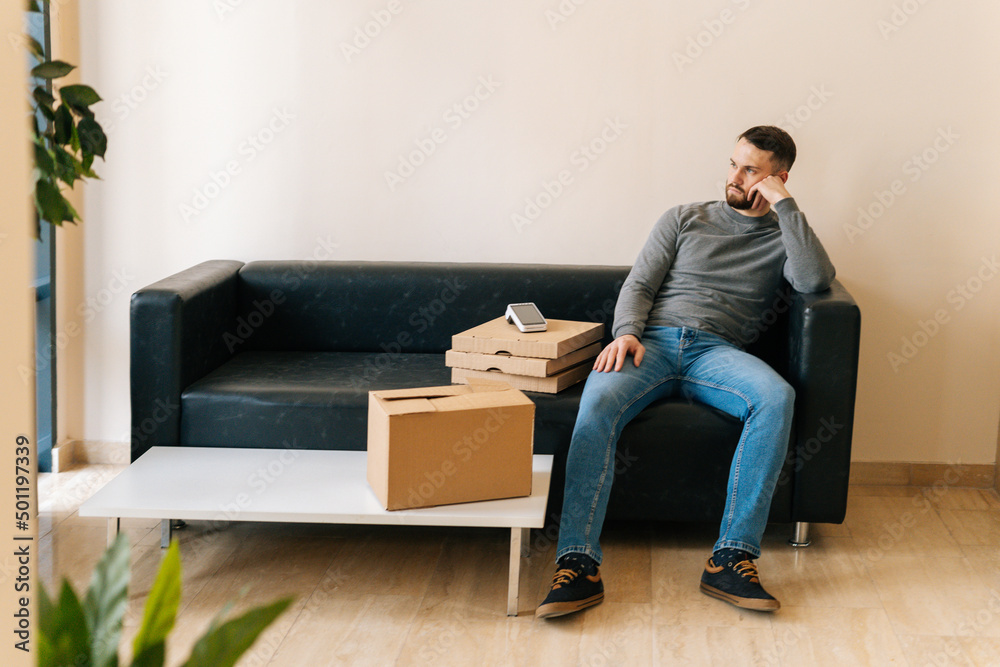 Portrait of delivery man waiting customer while sitting on sofa with boxes pizza, cardboard box and payment POS wireless terminal in hall of apartment or office building. Concept of online shopping.