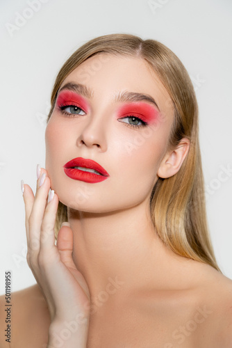 Model with naked shoulders and red makeup touching face isolated on grey.