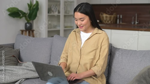 Asian young woman spending home leisure with laptop, working or studying remotel. Calm girl sitting at cozy sofa and looking at the laptop screen. Web browsing concept photo