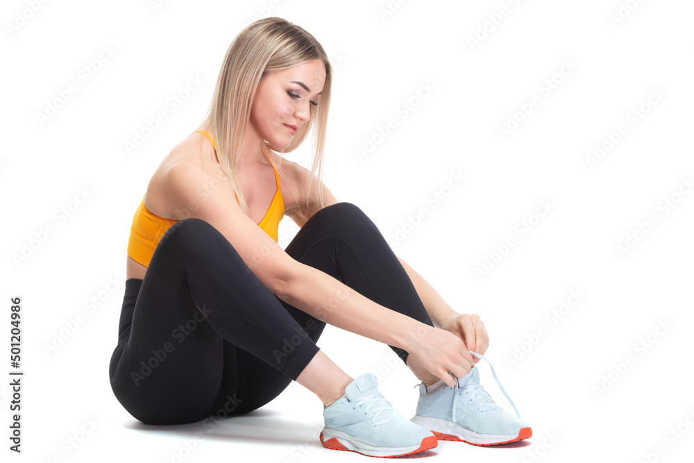 A beautiful, athletic, slim, smiling and cheerful woman in an orange top and black sweatpants is sitting and tying her shoelaces on sports sneakers. Lifestyle concept with sports and gym, healthy A beautiful, athletic, slim, smiling and cheerful woman in an orange top and black sweatpants is sitting and tying her shoelaces on sports sneakers. Lifestyle concept with sports and gym, healthy