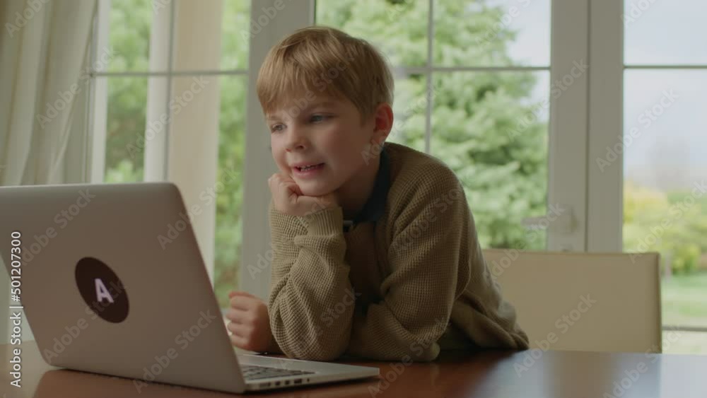 School boy talking using laptop sitting at the table by the window. Blonde kid learning online alone at home. School boy talking using laptop sitting at the table by the window. Blonde kid learning online alone at home.