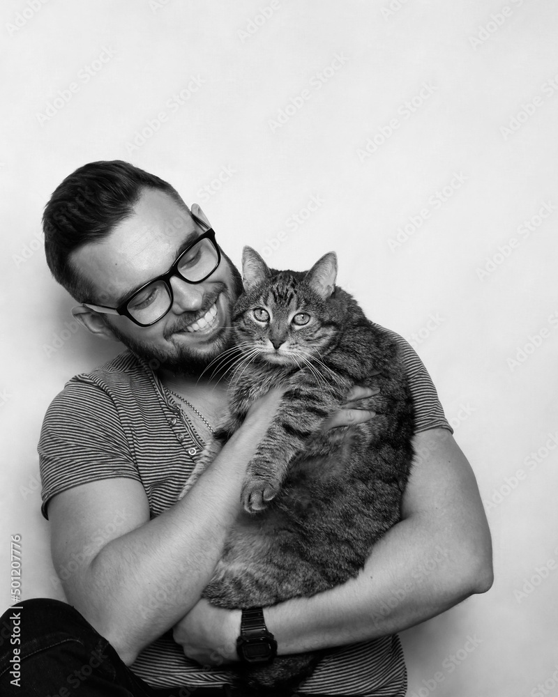 Studio portrait of a young smiling man with a beard and glasses holding a tabby cat in his arms. Grey background. White background. Animal love concept. Studio portrait of a young smiling man with a beard and glasses holding a tabby cat in his arms. Grey background. White background. Animal love concept.