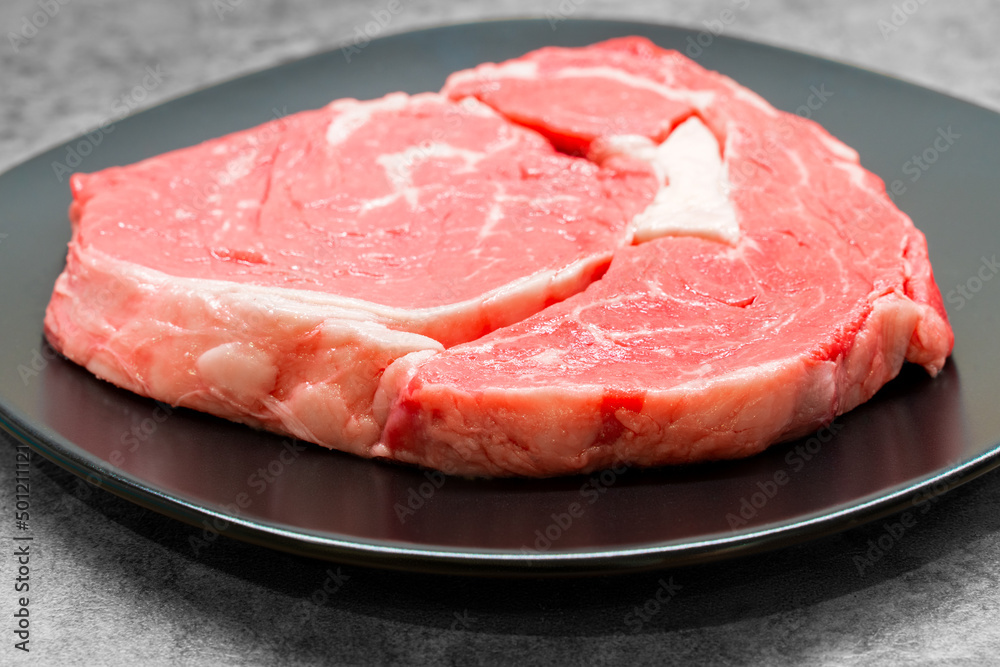 High angle studio shot of a raw beef rib eye steak on a black design plate standing on a grey concrete surface, with free copy space. Keto diet, warrior diet concept. Raw red meat ready for barbecue. High angle studio shot of a raw beef rib eye steak on a black design plate standing on a grey concrete surface, with free copy space. Keto diet, warrior diet concept. Raw red meat ready for barbecue.