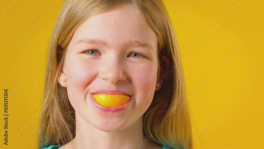 Studio Portrait Of Girl Using Orange Segment For Mouth And Teeth Against Yellow Background Studio Portrait Of Girl Using Orange Segment For Mouth And Teeth Against Yellow Background