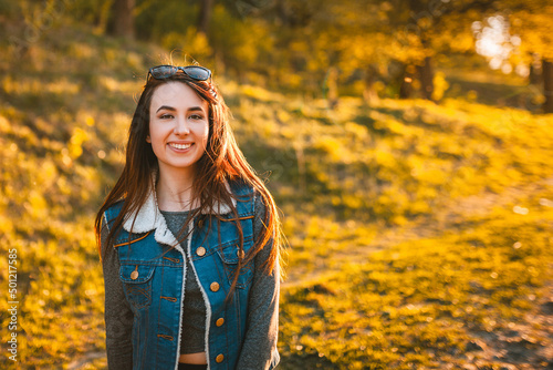 Beautiful girl in a blue denim jacket with glasses on her head stands at sunset. Spring sunbeams in the golden hour. The background is flooded with golden sunlight.