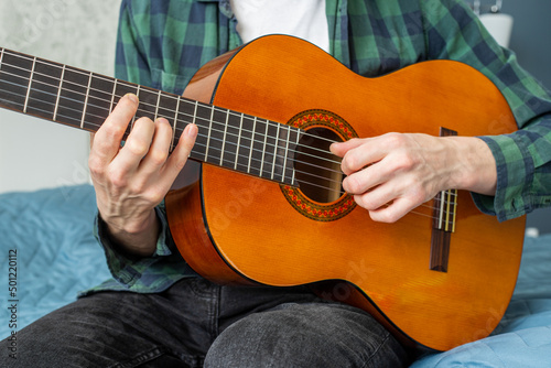 A man in a plaid shirt plays a natural-colored classical guitar sitting on a bed in close-up, selective focus.A male musician plays the guitar.Classical guitar in natural.Man playing acoustic guitar.