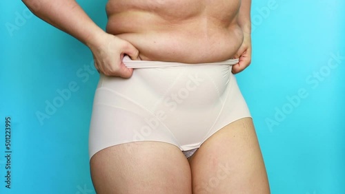 Unrecognizable fat plump overweight woman pulling on, adjusting beige seamless corrective bodysuit underwear shapewear on blue background. Slimming lingerie, body positive, obesity, weight loss, .