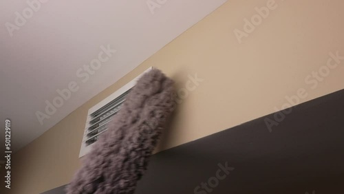 Small white rectangle furnace vent grill being dusted by a male hand with a long gray dusting wand. Wall air duct vent near a ceiling being dusted with a long gray dust wand by an adult male hand.