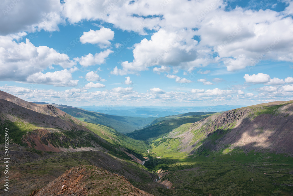 Naklejka premium Scenic aerial view from mountain pass to green forest valley among mountain ranges and hills on horizon at changeable weather. Green landscape with sunlit mountains under cumulus clouds in blue sky.
