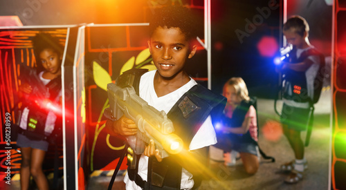 Portrait of happy cheerful tweenager boy with laser gun having fun on dark lasertag arena