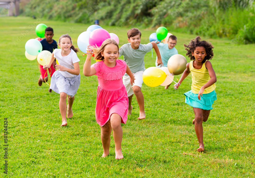 Happy tween friends of different nationalities having fun together in summer city park, chasing each other on green meadow, holding colorful balloons in hands..