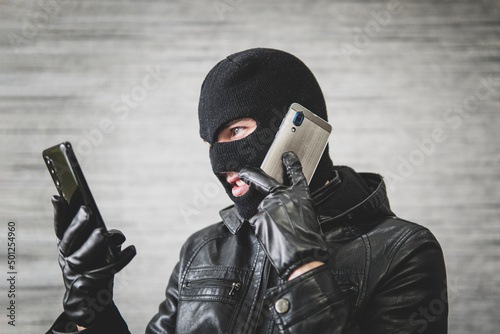 Young adult in black clothes with hidden face. Ill-intended fraudster uses mobile. Fraudster calls. Scam. Mobile racket. Hacker hijacks by phone. Cellphone account fraud.