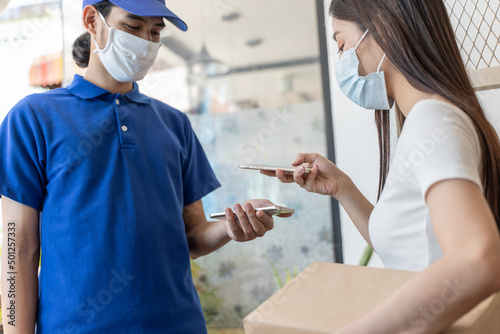 Young Asian woman wearing face mask using smartphone for scan QR code accept delivery package from delivery staff for prevent covid-19 infection during coronavirus pandemic.