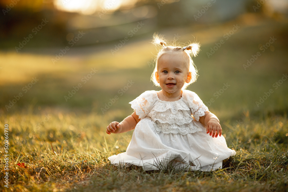 Happy little girl sits on the grass outdoors. Cute baby girl with blond hair is walking in park. Adorable kid play with grass. concept of happy childhood, games, fun. Family leisure. outdoor play