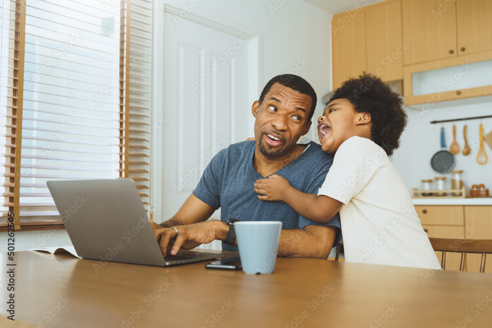African American father working from home on laptop while his Cheerful son distracts him in kitchen. African American father working from home on laptop while his Cheerful son distracts him in kitchen.