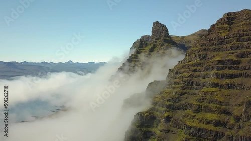 Aerial rising over verdant steep hill summit, clouds covering mountains in background, in Djúpivogur small town near Iceland fiords at daytime photo