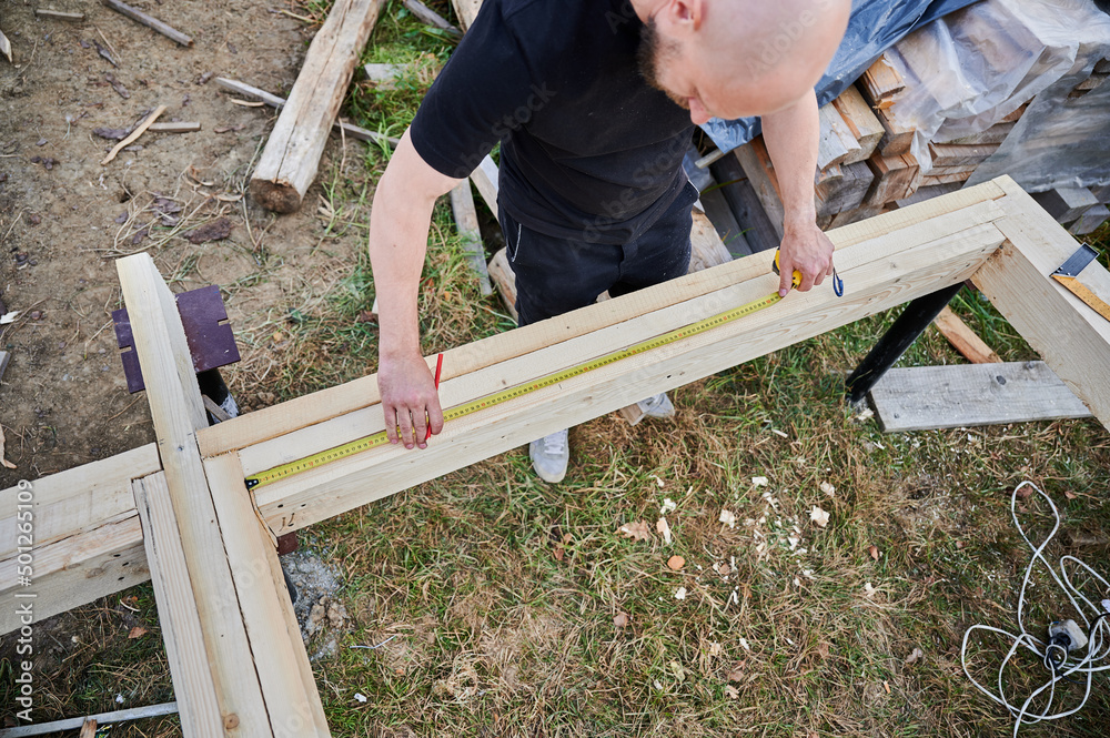 Man worker building wooden frame house on pile foundation. Carpenter using tape measure for measuring wooden planks and making marks with pencil. Carpentry concept. Man worker building wooden frame house on pile foundation. Carpenter using tape measure for measuring wooden planks and making marks with pencil. Carpentry concept.