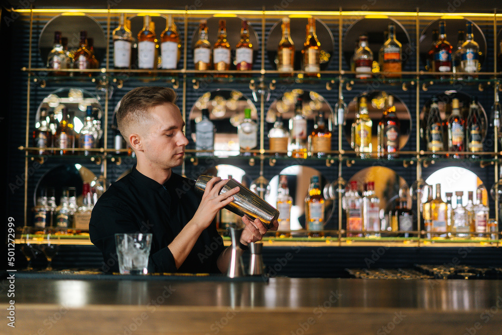 Front view of focused bartender mixing ingredients of fresh cocktail by shaking shaker standing behind bar counter, on blurred background of shelves with different alcoholic drinks, selective focus.