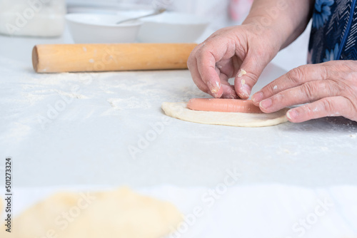 Senior woman hands making pies with sausage on a white kitchen table with wooden rolling pin on background. Selective focus. Cooking at home concept. Tradition home-made food