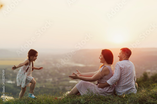 Happy family concept. Young happy mother, father and their little cute daughter having fun together on the mountain at sunset. Daughter running to the parents.