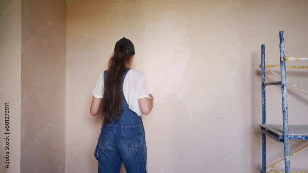 Professional painter female paints the wall with roller and tray on construction site. Young woman in overalls and cap doing home decoration and renovetion. Worker makes DIY repair work in the room. Professional painter female paints the wall with roller and tray on construction site. Young woman in overalls and cap doing home decoration and renovetion. Worker makes DIY repair work in the room.