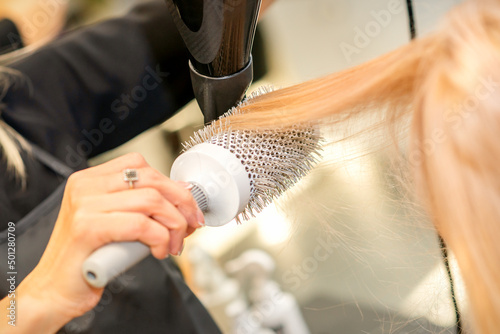 Drying straight blond hair with black hairdryer and white round brush in hairdresser salon, close up