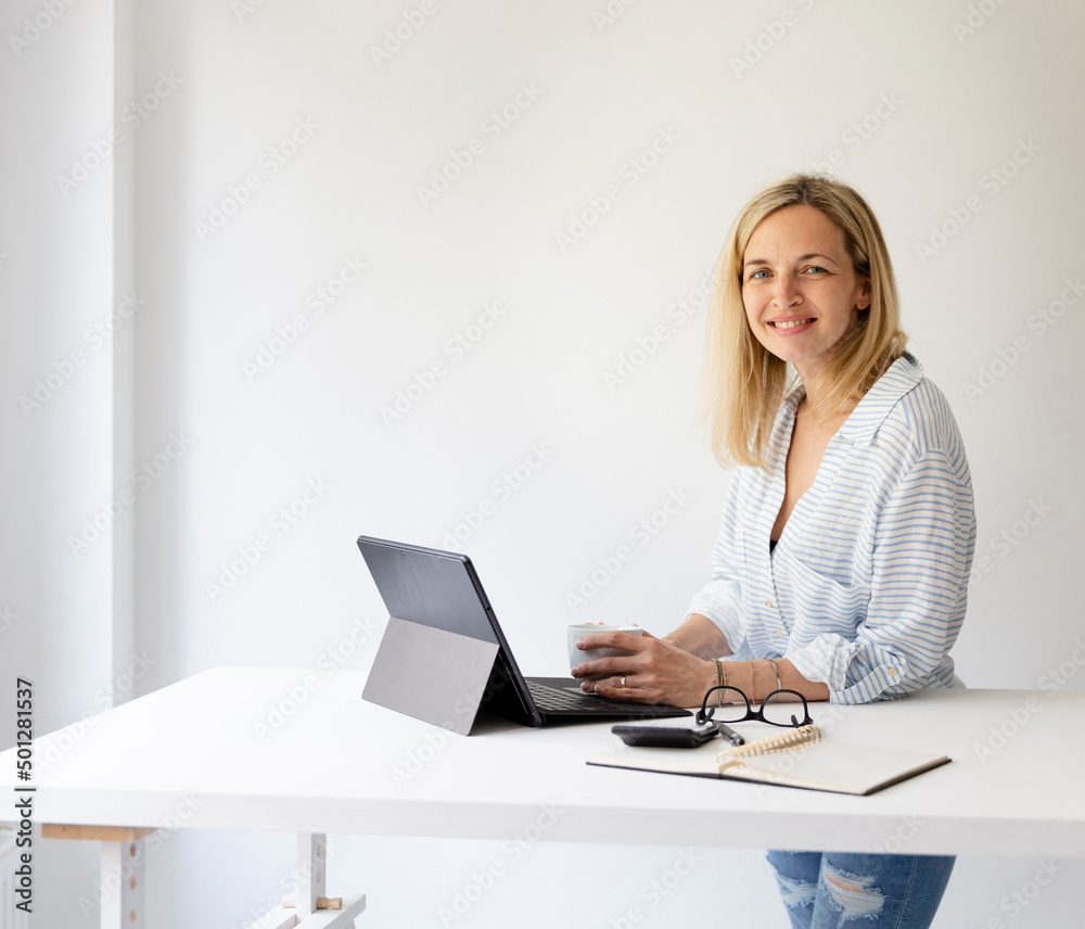 pretty young blonde business woman stands by her ergonomic height adjustable work desk and works on tablet from home in her home office pretty young blonde business woman stands by her ergonomic height adjustable work desk and works on tablet from home in her home office