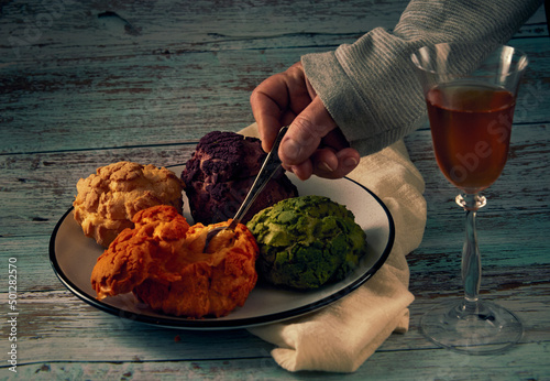 Several multi-colored profiteroles of cakes on a ceramic plate, a hand with a teaspoon and a glass of misted white wine on a wooden table photo