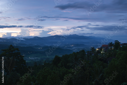 Indonesian tender landscape - sunrise with fresh morning sky, light pink clouds, sunbeams over hazy blue mountains, green valley, tropical forest, houses with yellow lights on Bali, Munduk village.