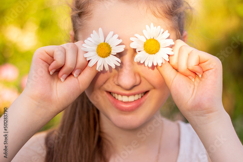 A beautiful young girl smiles at the camera and holds flowers chamomile in her hands