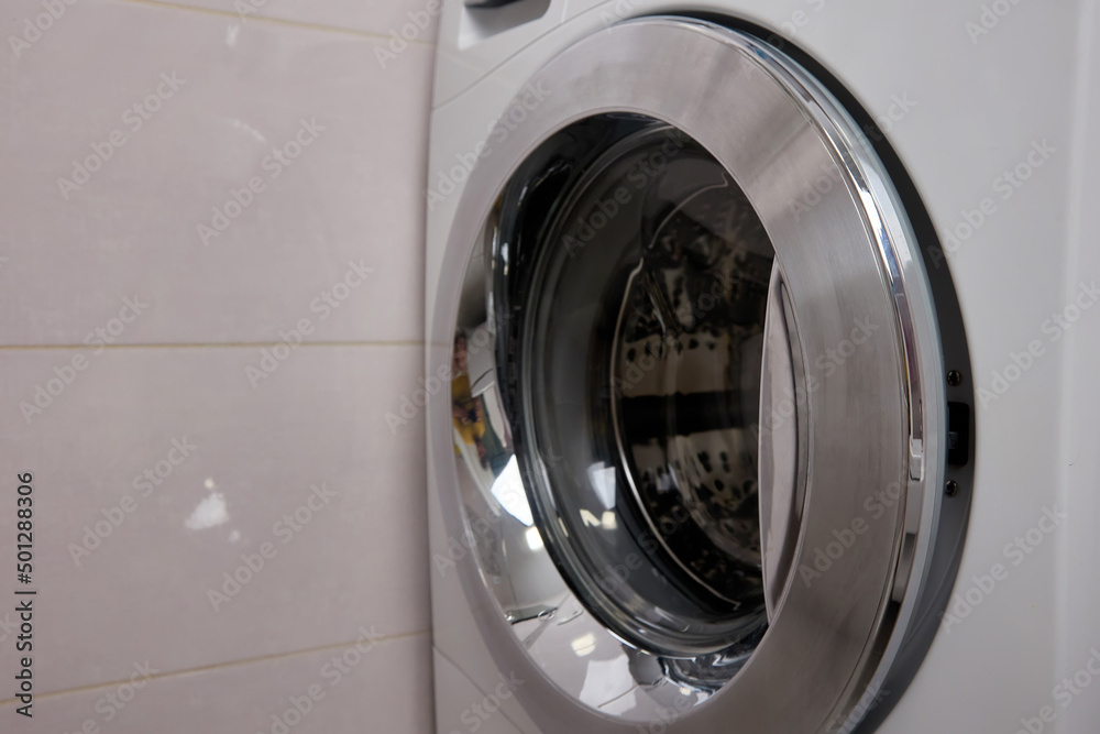 A modern washing machine with a metal door standing near a tiled wall in the room, a place for text. Bathroom interior A modern washing machine with a metal door standing near a tiled wall in the room, a place for text. Bathroom interior