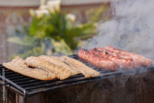 Man cooking botifarra sausage and bacon on the barbecue grill for an outdoor summer party. Barbecue smoke. Background of food.