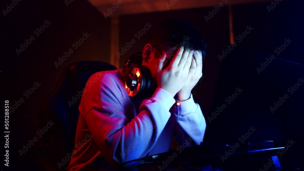 Man is crying and upset after losing an online videogame on laptop, covers face with hands, front view. Young man cries after losing in videogame