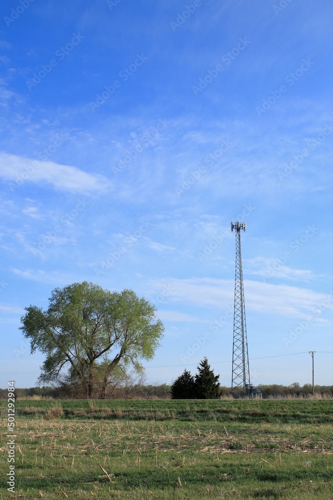A 5G cell phone tower east of Nickerson Kansas USA with blue sky and clouds out in the country with green trees.