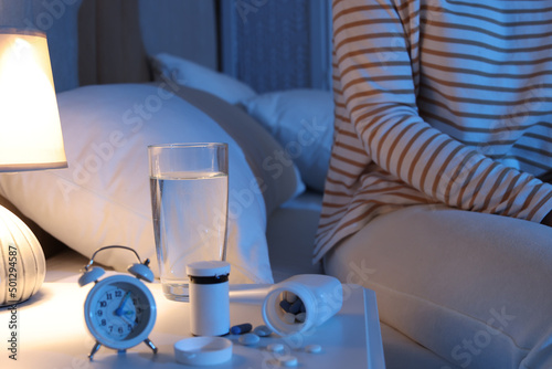 Woman near nightstand with pills, water and alarm clock in bedroom at night, closeup